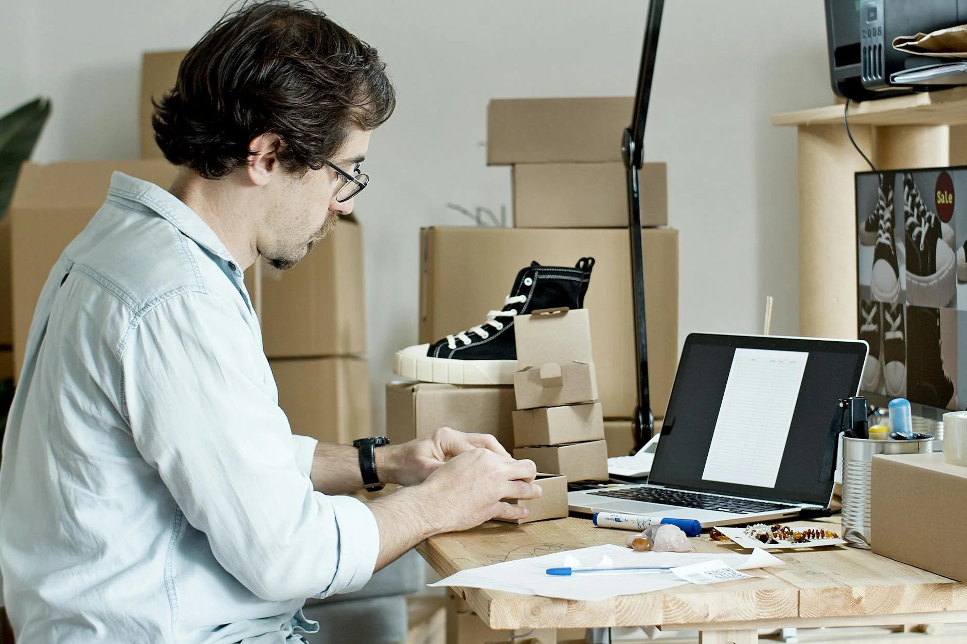 Persona en un pequeño taller doméstico empaquetando un producto artesanal sobre una mesa de madera, con etiquetas adhesivas y un portátil al lado